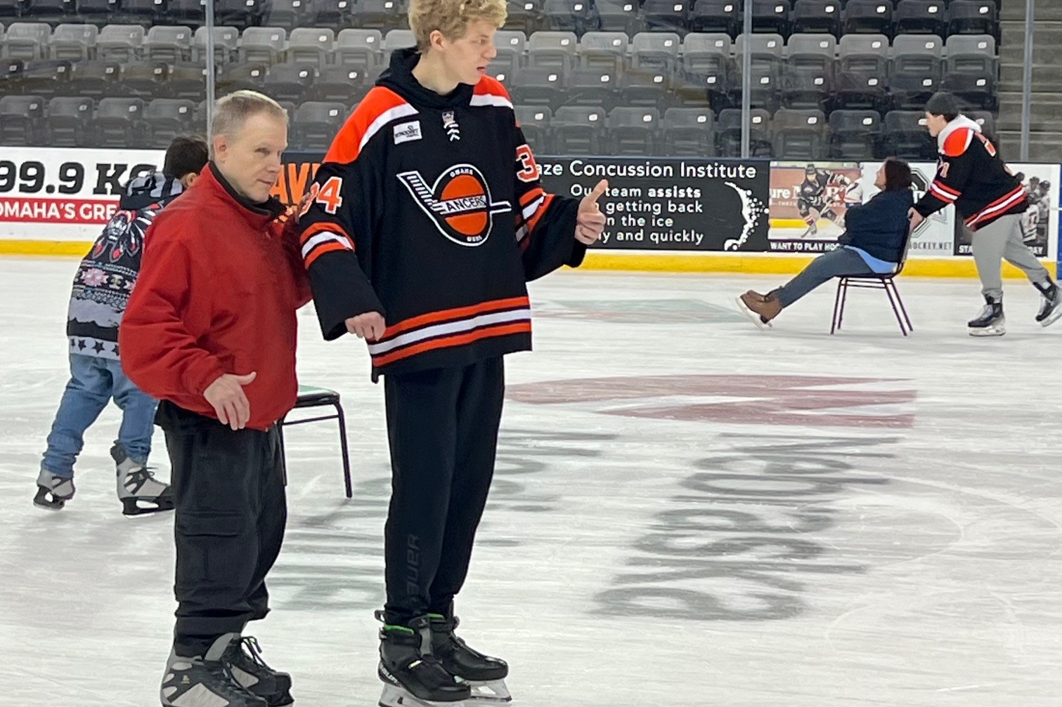Tony, a man with gray hair is holding onto a Lancer players arn, using him as a sighted guide while ice skating. There are other people ice skating in the background at an indoor ice rink.