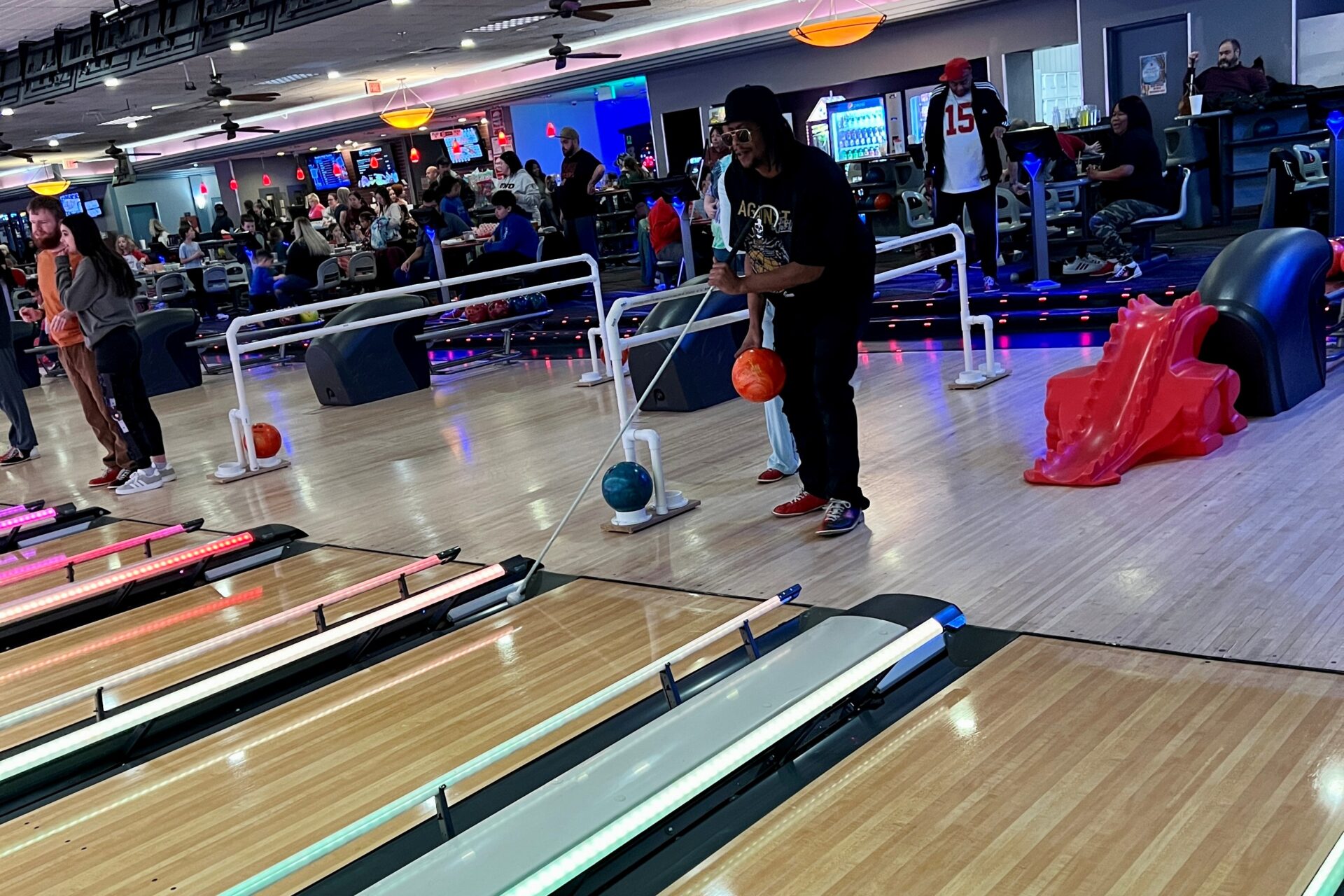 Mortel wearing sunglasses has a bowling ball in one hand and is his white cane in the other hand as he steps up to a bowling lane.