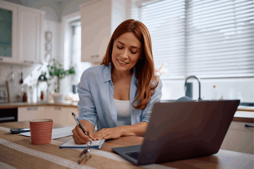 A woman is sitting down, taking notes with her computer in front of her.