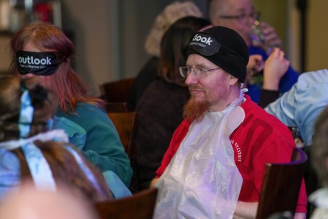 Dustin wearing a bib and his sleep shades on his forehead is sitting next to a woman with her sleep shades on.