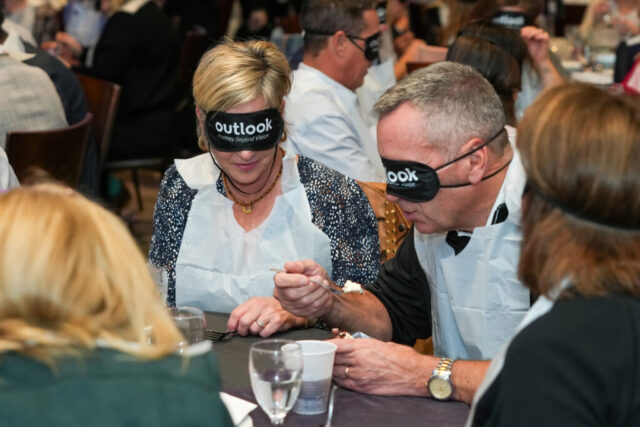 A woman is wearing sleep shades and holding her fork while the man next to her is mid bite and wearing sleep shades.