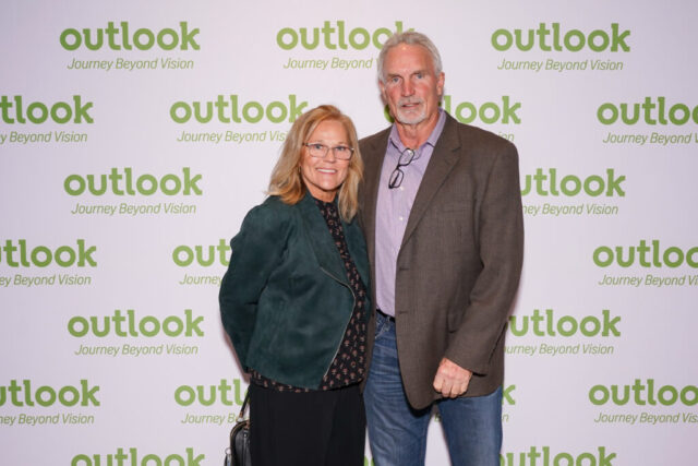 A man and woman smiling in front of an Outlook backdrop.