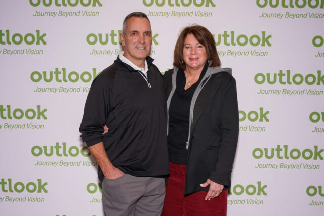 A man and woman smiling in front of an Outlook backdrop.