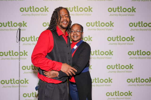 Mortel and a woman are smiling and hugging in front of an Outlook backdrop.