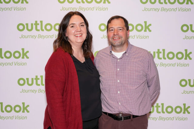 A man and woman smiling in front of an Outlook backdrop.