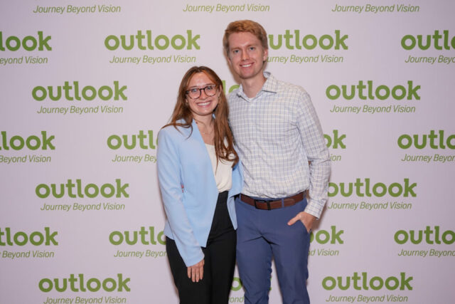 A man and woman smiling in front of an Outlook backdrop.