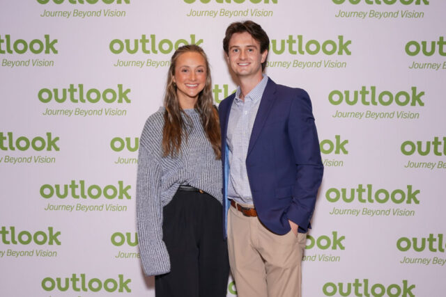 A man and woman smiling in front of an Outlook backdrop.