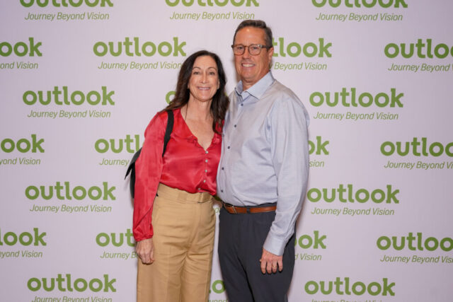 A man and woman smiling in front of an Outlook backdrop.