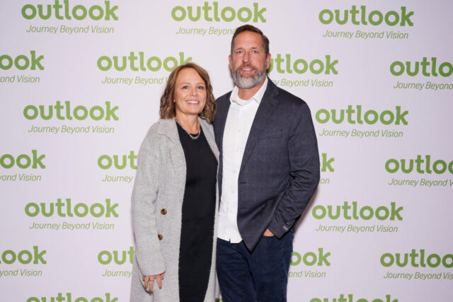 A man and woman smiling in front of an Outlook backdrop.