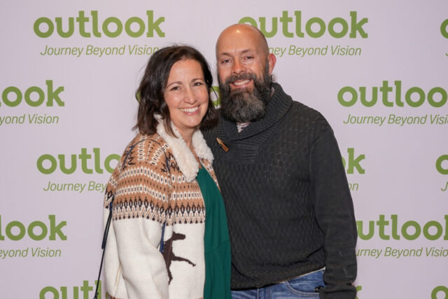 A man and woman smiling in front of an Outlook backdrop.