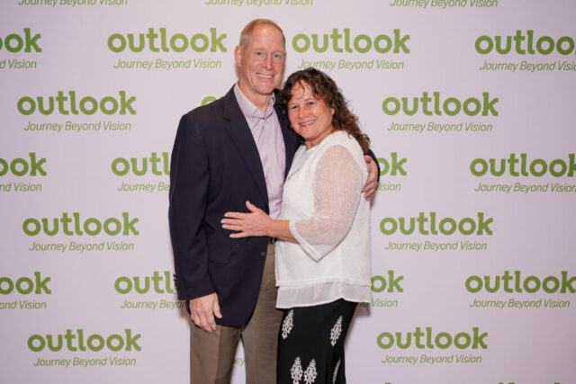A man and woman smiling in front of an Outlook backdrop.