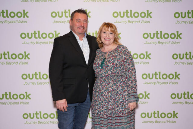 A man and woman smiling in front of an Outlook backdrop.