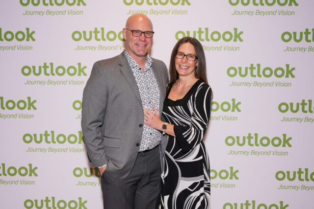 A man and woman smiling in front of an Outlook backdrop.