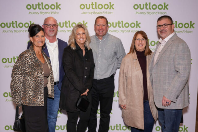 Three pairs of a man and woman are smiling in front of an Outlook backdrop.