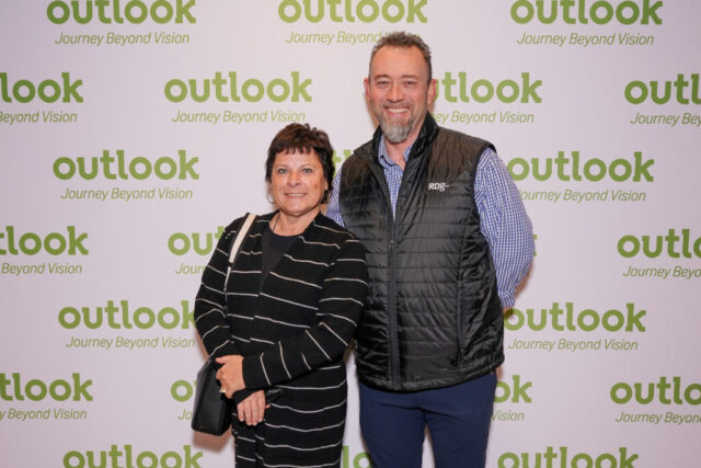 A man and woman smiling in front of an Outlook backdrop.