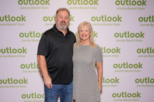 Stephanie and husband are smiling in front of an Outlook backdrop.