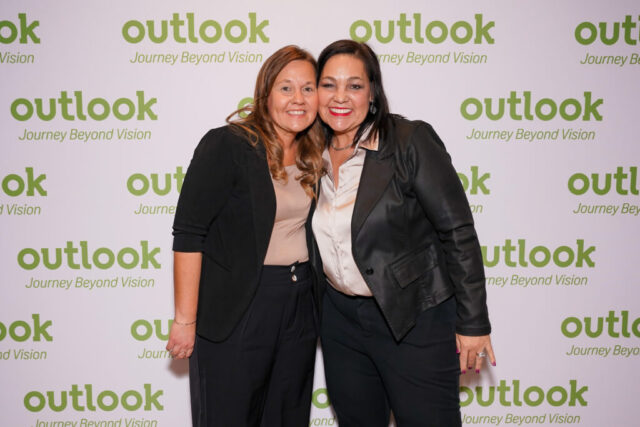 Two women smiling in front of an Outlook backdrop.