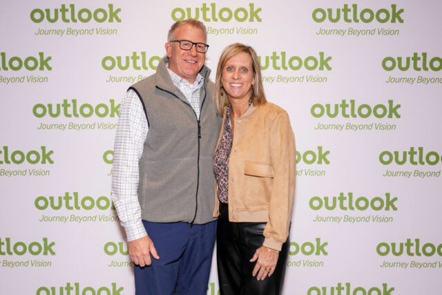 A man and woman smiling in front of an Outlook backdrop.