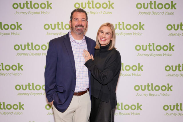 Tom and his wife are smiling in front of an Outlook backdrop.