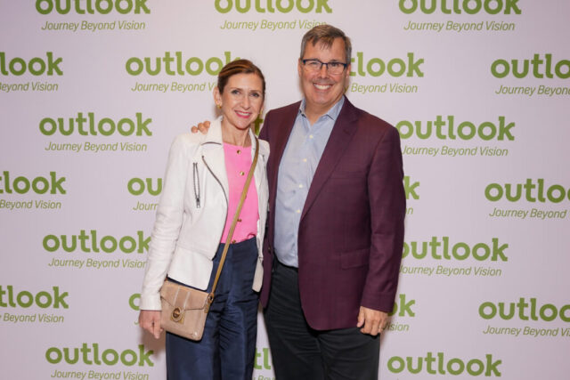 A man and woman smiling in front of an Outlook backdrop.