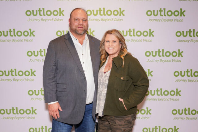 A man and woman smiling in front of an Outlook backdrop.
