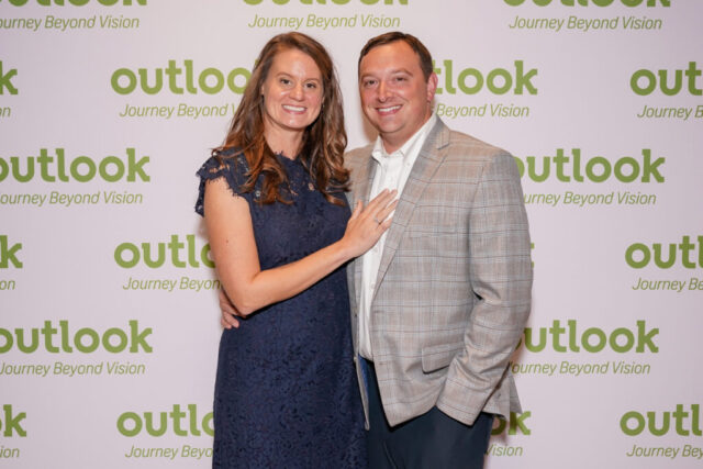 Ashley and her husband are smiling in front of an Outlook backdrop.