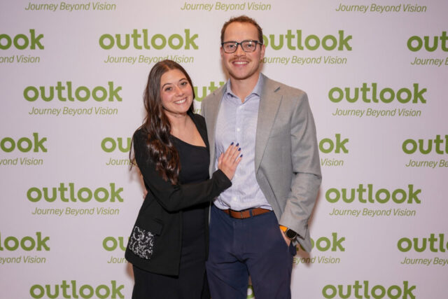 A man and woman smiling in front of an Outlook backdrop.