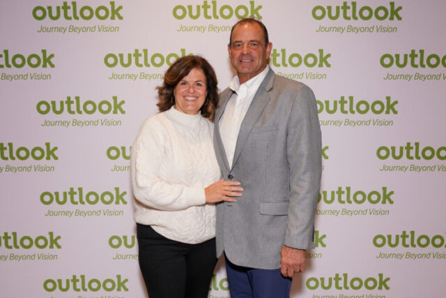 A man and woman smiling in front of an Outlook backdrop.