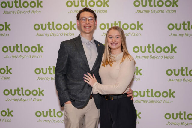 A man and woman smiling in front of an Outlook backdrop.