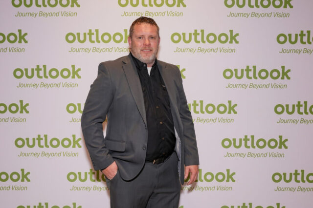 A man is smiling in front of an Outlook backdrop.