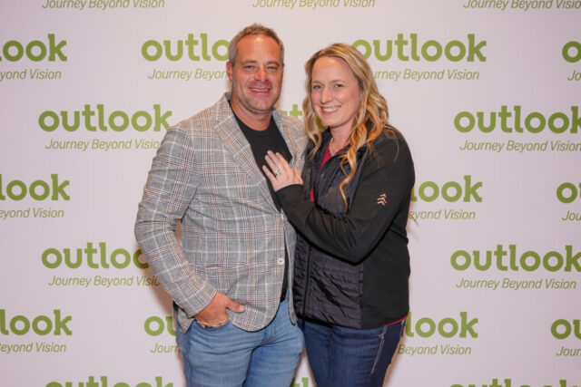 A man and woman smiling in front of an Outlook backdrop.