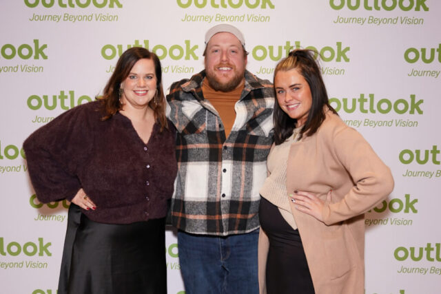 Ashley, Meghann, and a man are smiling in front of an Outlook backdrop.