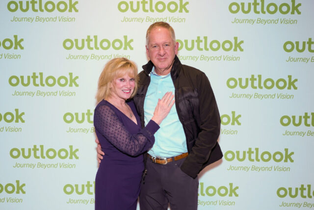 A man and woman smiling in front of an Outlook backdrop.