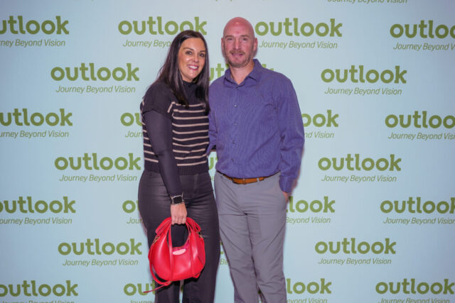 A man and woman smiling in front of an Outlook backdrop.