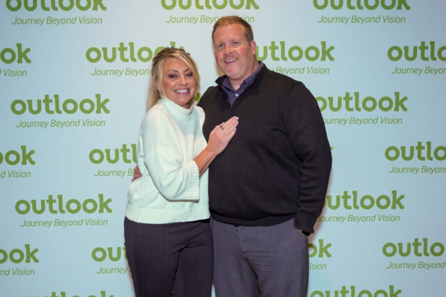 A man and woman smiling in front of an Outlook backdrop.