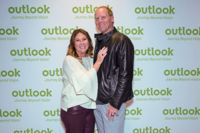 A man and woman smiling in front of an Outlook backdrop.