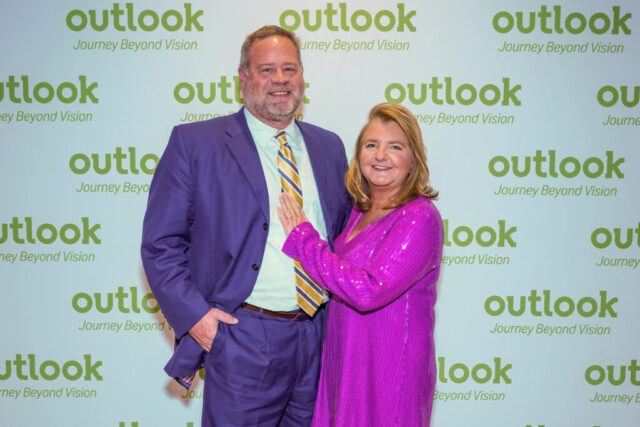 A man and woman smiling in front of an Outlook backdrop.