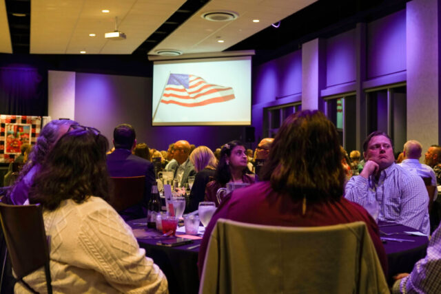 A video is being played in the background with an American flag on the screen. Table guests are watching.