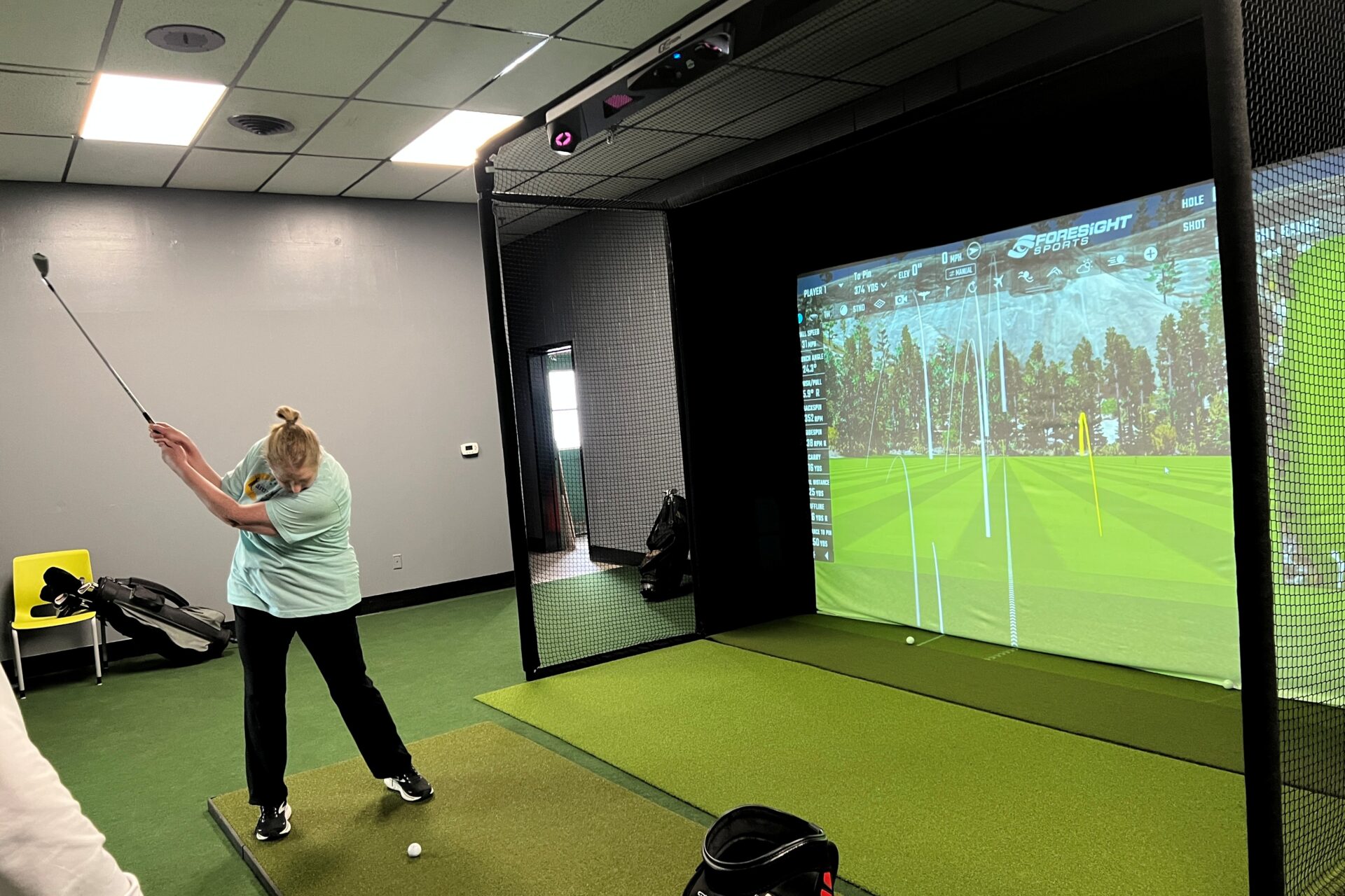 Kristal looking down while swinging a golf club aimed at a large simulator screen of a golf course.
