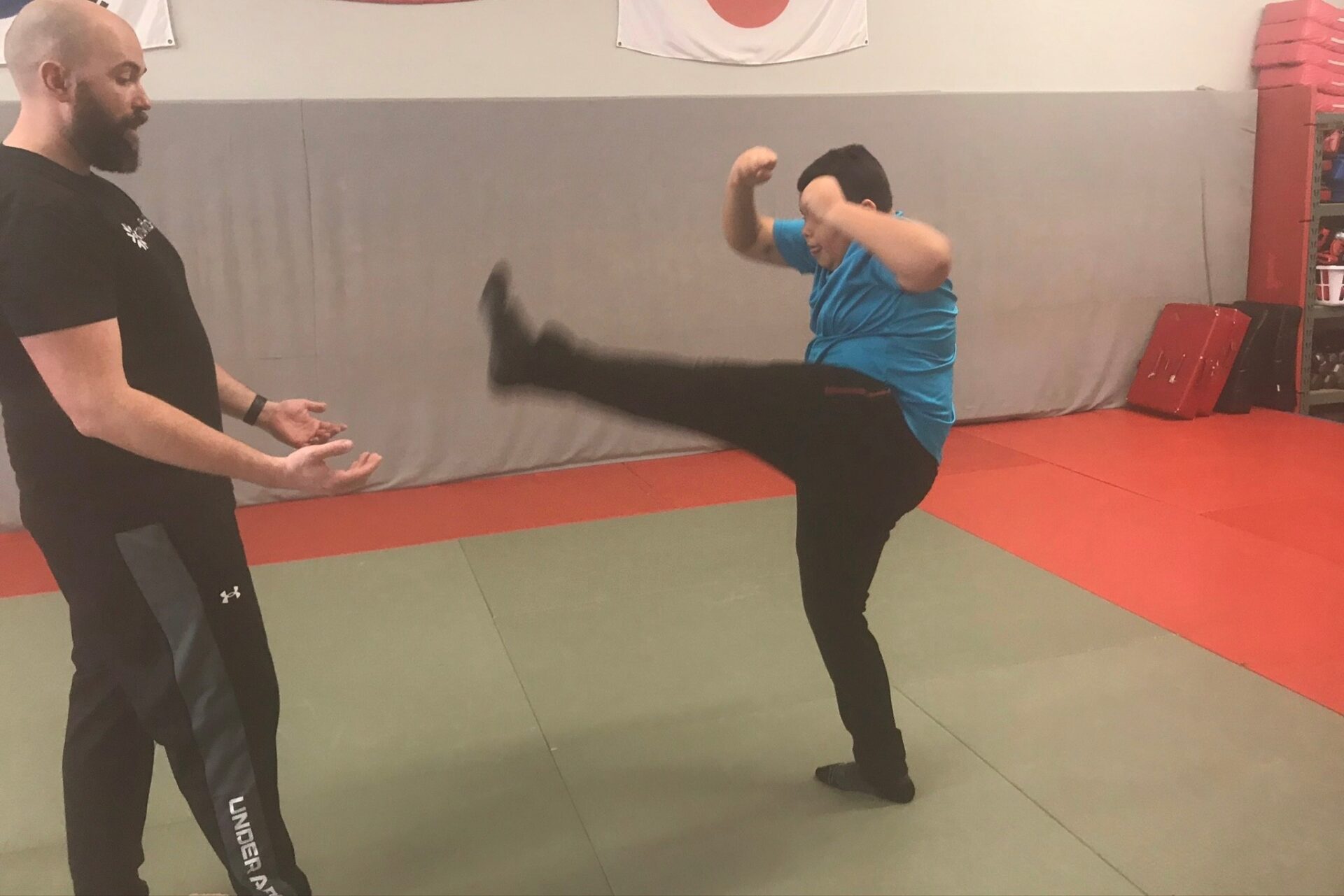 A young man with his fists clenched, blocking his face and his leg kicking out in front of him. He is receiving directions from an instructor.