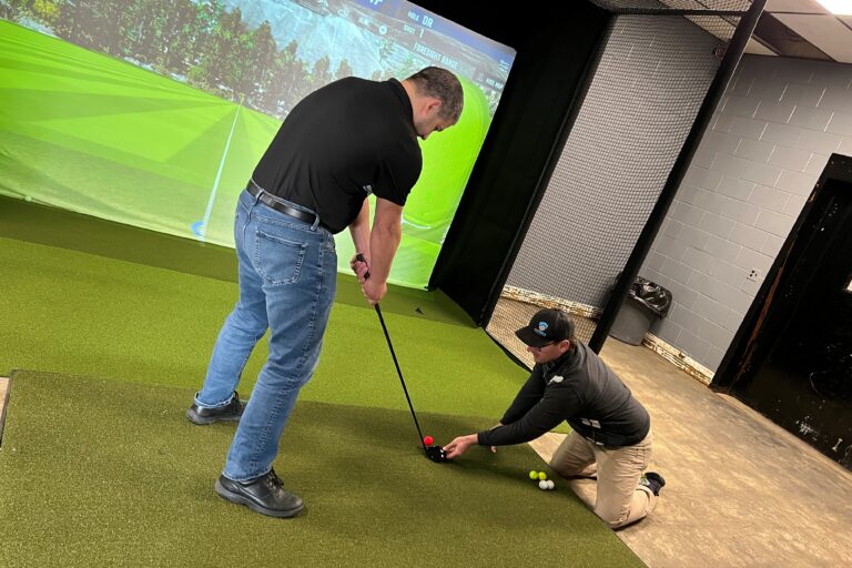 Ross, a visually impaired golfer holds a golf club, while volunteer Steve kneels down to line up a golf ball near Ross's golf club.