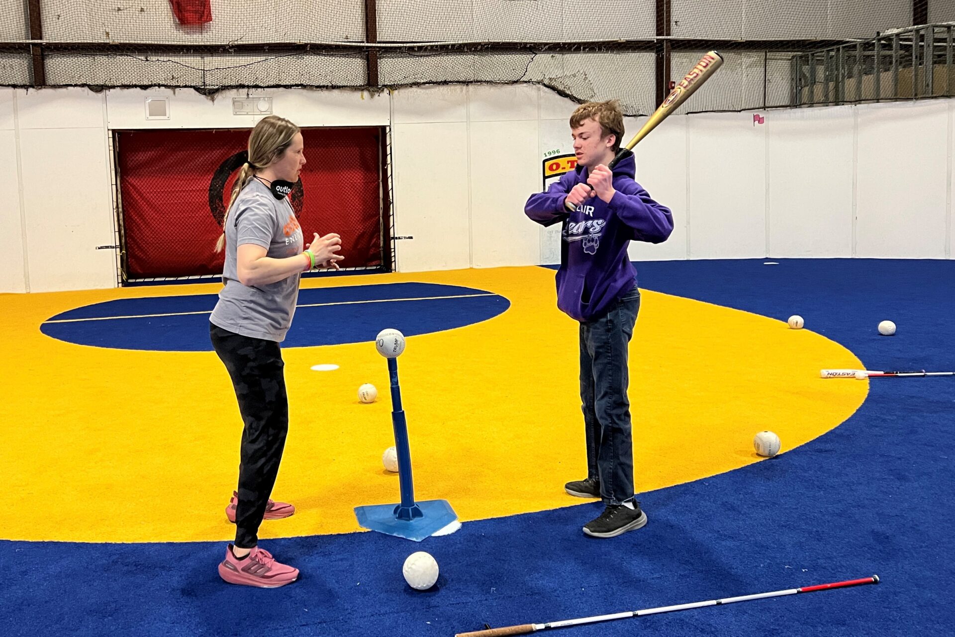 A teenager, Andrew, and a volunteer standing face to face between them is a batting tee with a beeping ball. Andrew is getting ready to swing a bat.