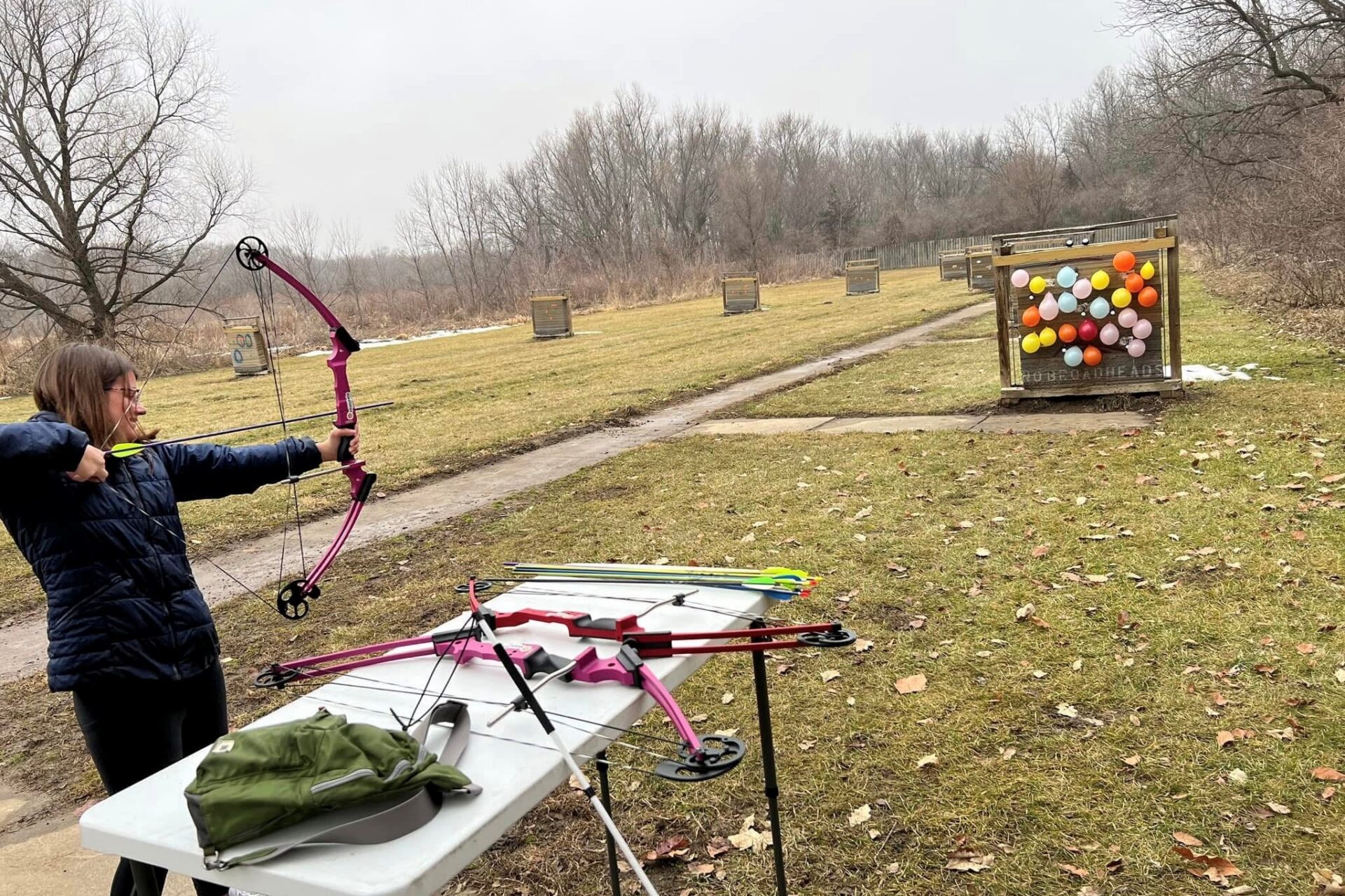 Camille is aiming a bow and arrow at a target covered with a bunch of small balloons. She is outside with the woods of Lake Cunningham in the background.
