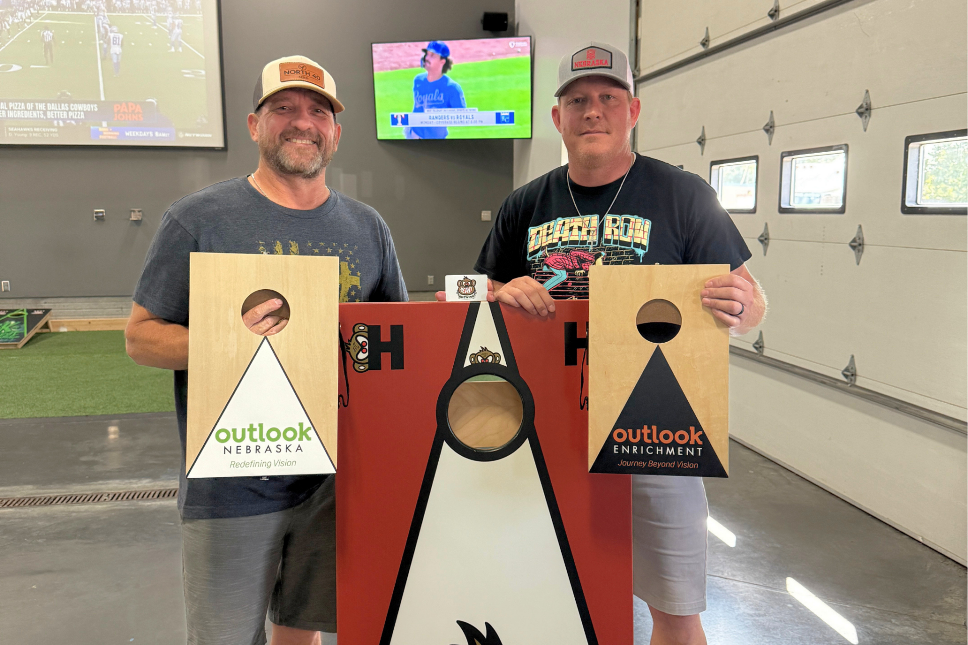 Two men are smiling between a Heavy Brewing cornhole board, and each is holding an Outlook logoed mini cornhole board.