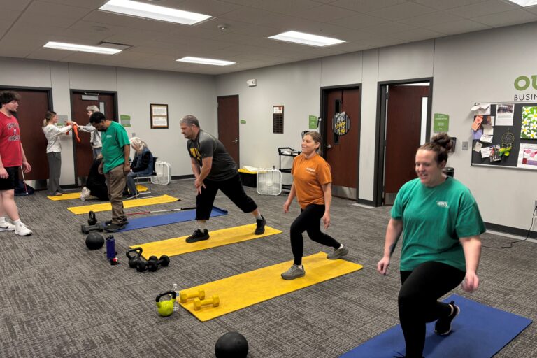 A row of people lunging on yoga mats. One woman is doing exercises while sitting on a chair.