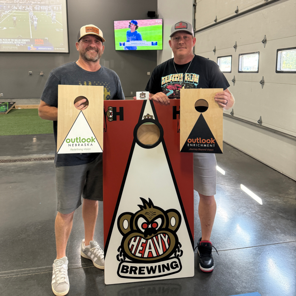 Cornhole tournament participants with Outlook Enrichment branded cornhole boards and Heavy Brewing logo.