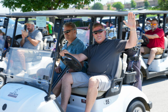 Two men in their golf cart. The one on the right is waving at the camera.