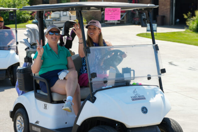 Two ladies from Gallagher on their golf cart. One is giving a piece sign and the other is waving.