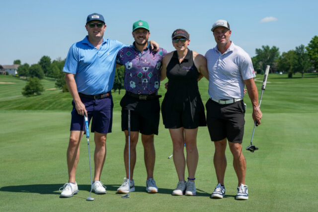 A group of three men and one woman smiling on the green.