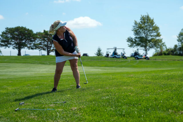 A lady is swinging her club on the green. Golf carts are in the background.